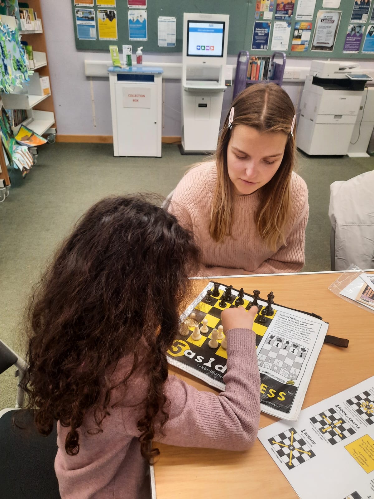 A woman teaching a young girl to play 5asideCHESS in a library