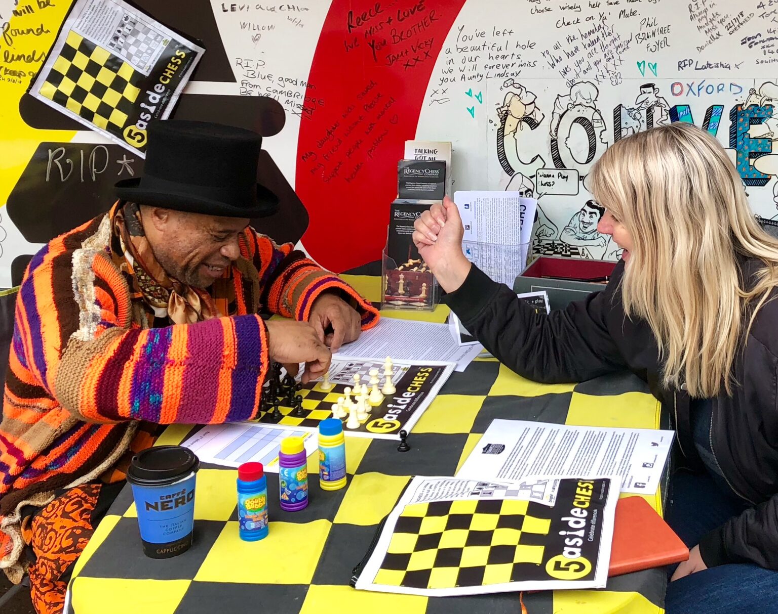 Two strangers laughing over a 5asideCHESS board at a street table in Oxford