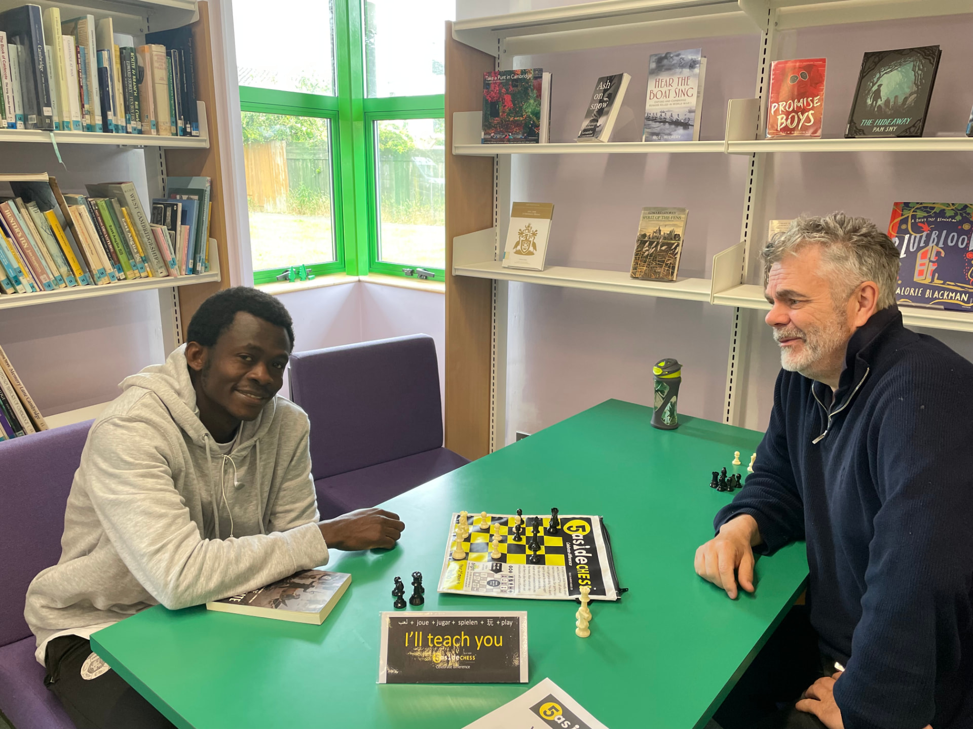 A man teaching another how to play 5asideCHESS across a green library table